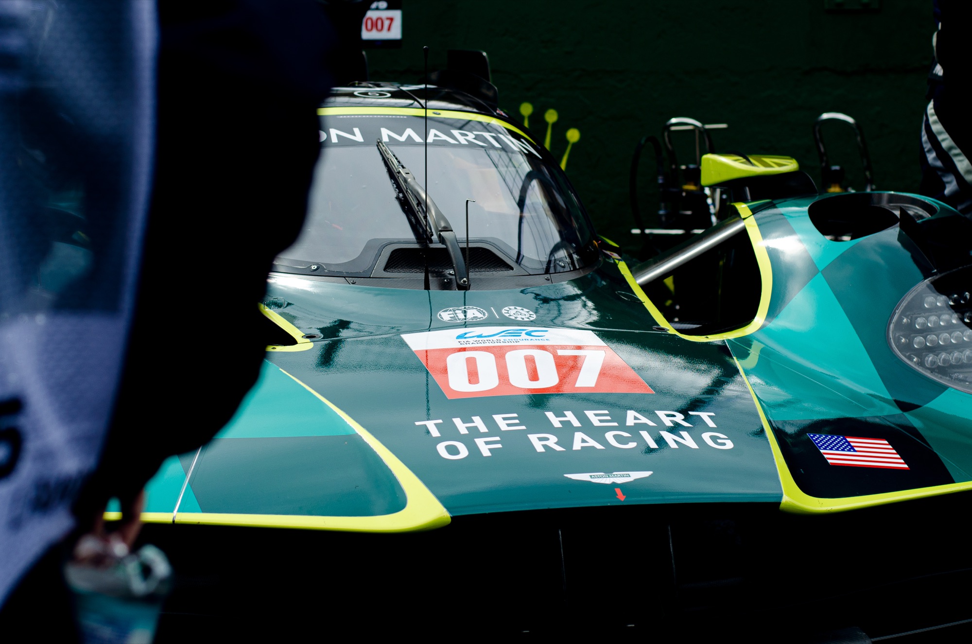 Aston Martin Valkyrie #007 da The Heart of Racing parado no box, alguém passando em primeiro plano