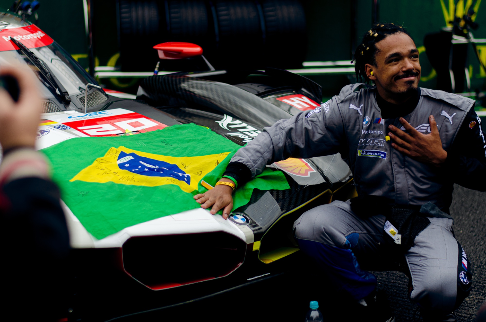 Membro da BMW M Team WRT ajoelhado ao lado do BMW M Hybrid V8 #20, com a bandeira do Brasil autografada sobre o bico