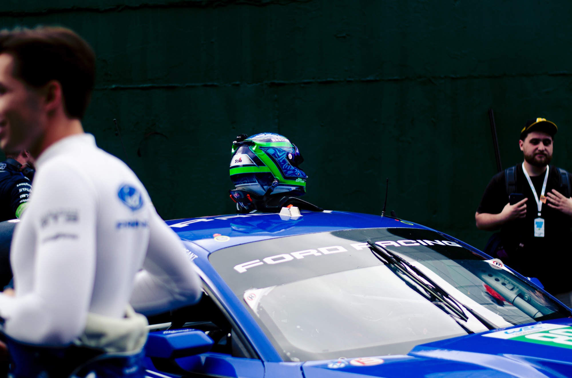 Capacete apoiado no teto do Ford Mustang GT3 azul, piloto de nomex branco desfocado em primeiro plano